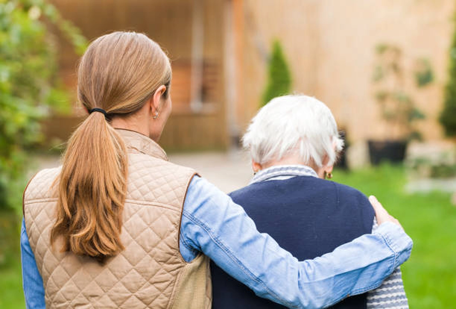Adler Pflegedienst - Pflege und Unterstützung älterer Menschen - Eine Frau mit blondem Pferdeschwanz ist von hinten zu sehen, wie sie geht und eine ältere Person mit dem Arm um sie stützt.