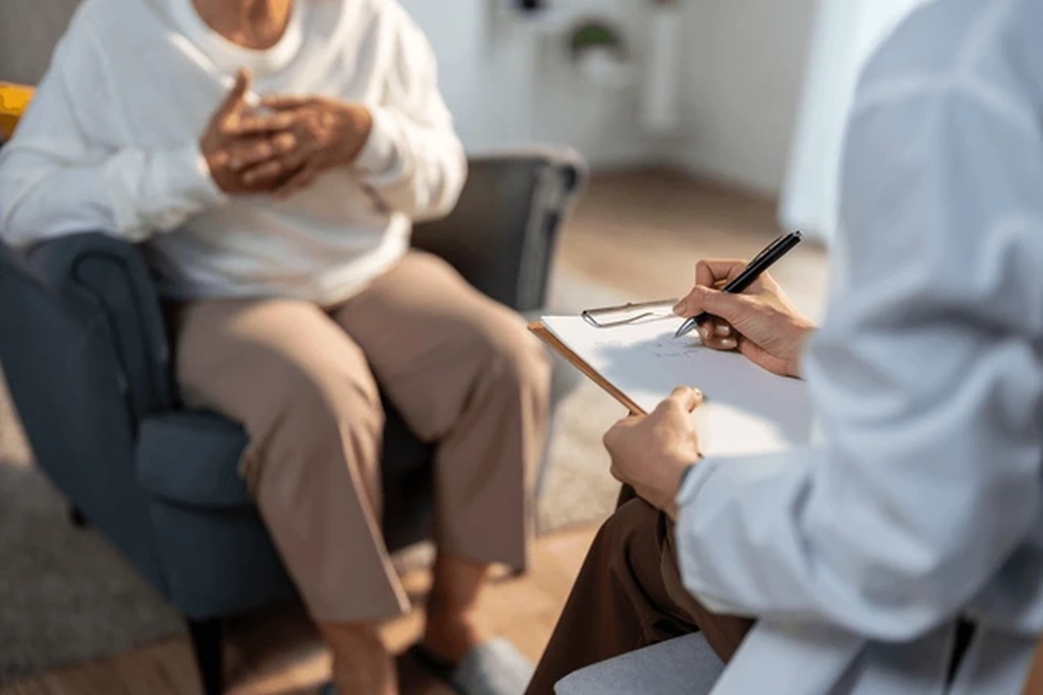 Adler Pflegedienst - Pflege und Unterstützung älterer Menschen - Eine über die Schulter blickende Aufnahme zeigt einen Angehörigen der Gesundheitsberufe oder einen Therapeuten, der mit Klemmbrett und Stift in der Hand Notizen macht, während er einer Person auf einem Stuhl gegenübersitzt.